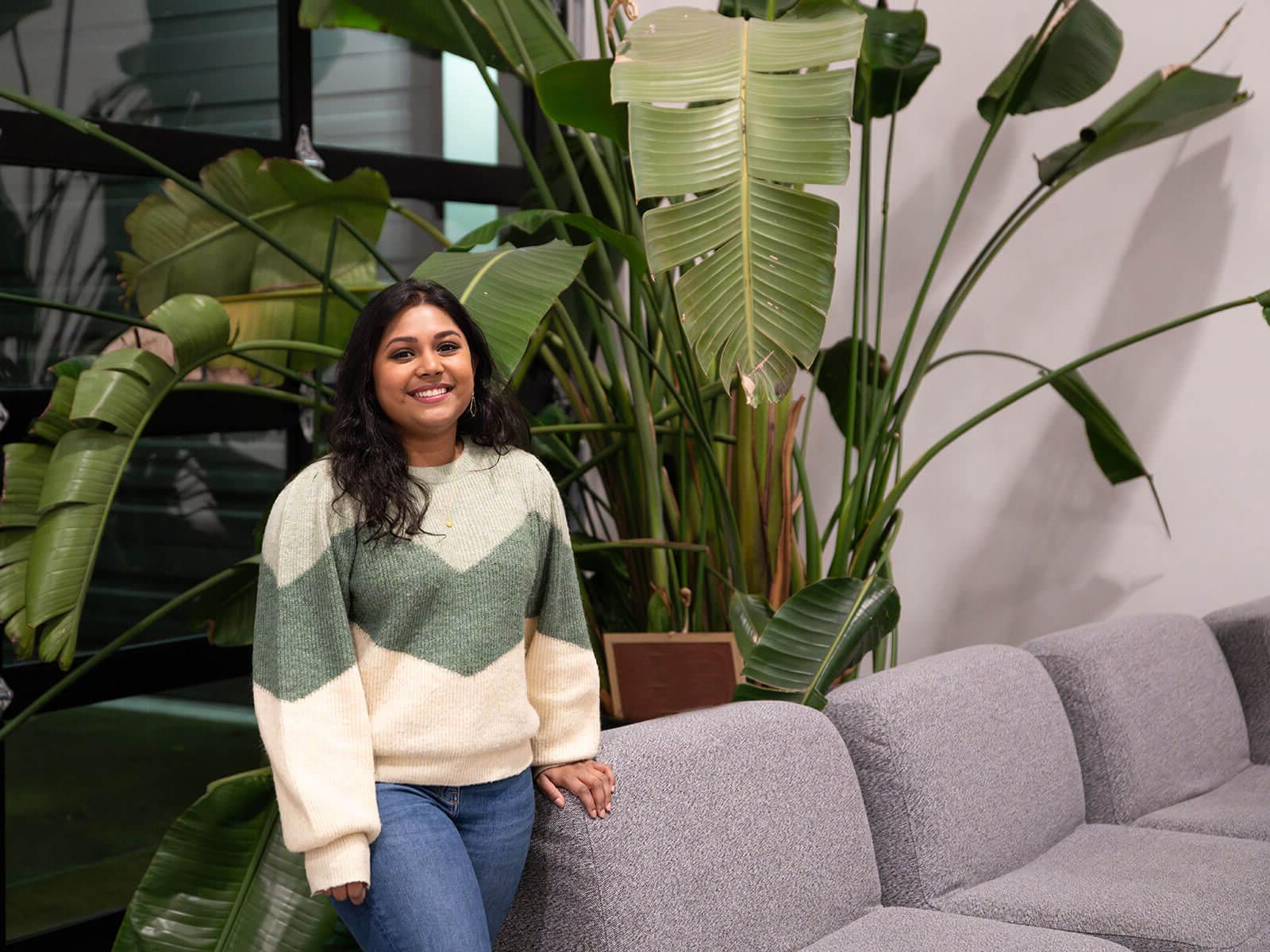 DigiPen graduate Neha Chintala smiles standing next to a couch and some large, leafy plants.